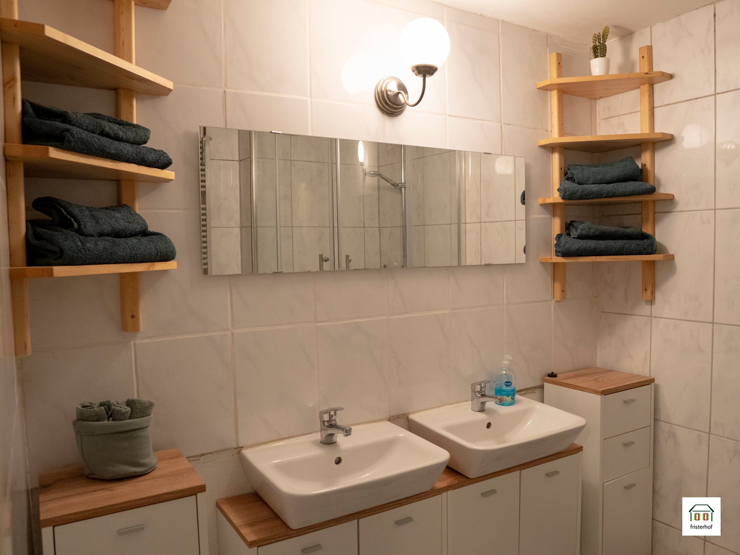 Badezimmer mit zwei Waschbecken unter einem breiten Spiegel und Holzregalen mit Handtüchern