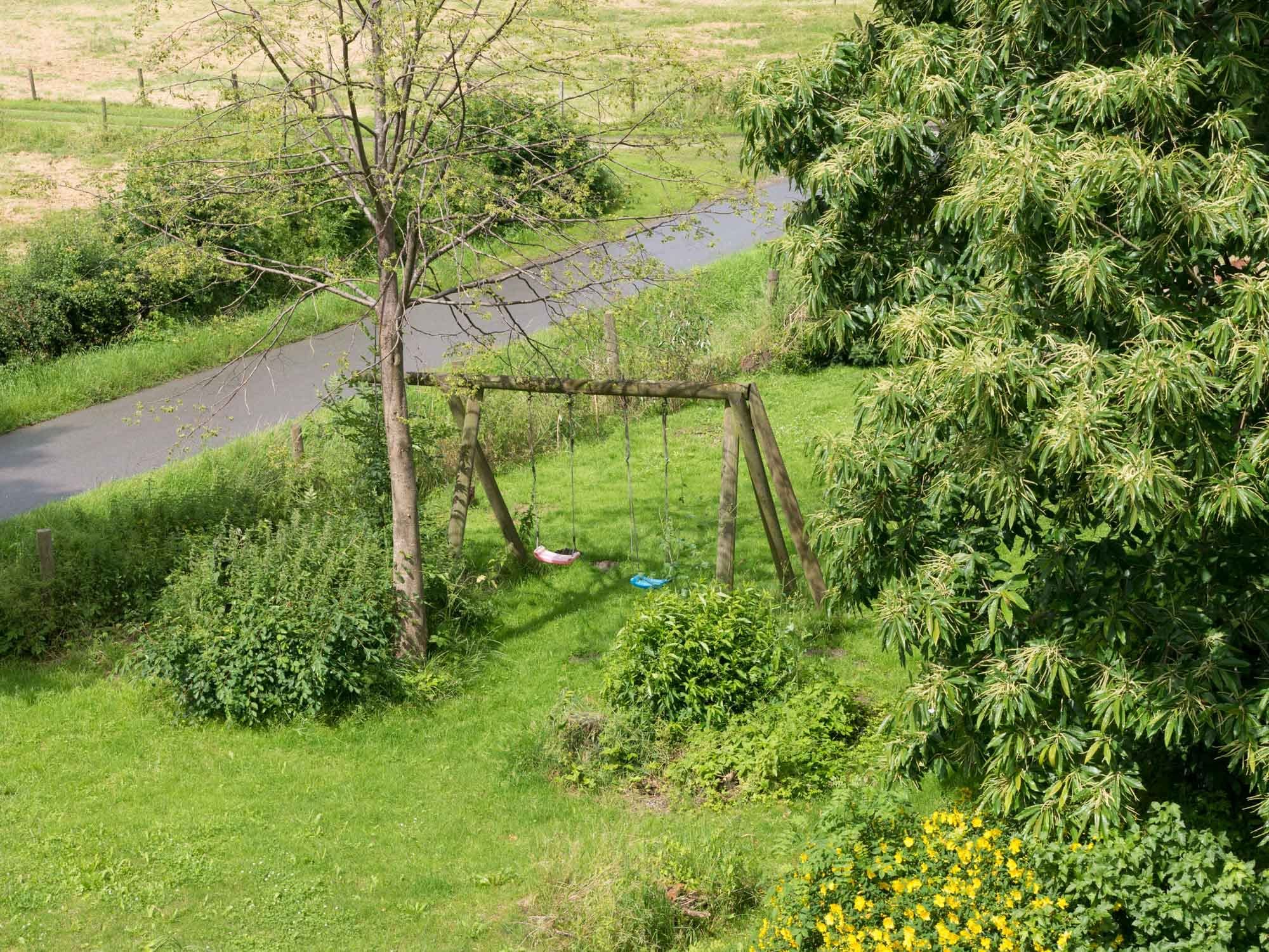 Holzschaukeln auf dem Rasen zwischen den Bäumen, dahinter die Deichstraße